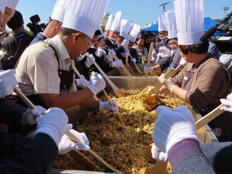 흰 모자를 쓴 사람들이 큰 솥에서 송이 비빔밥을 함께 비비는 모습