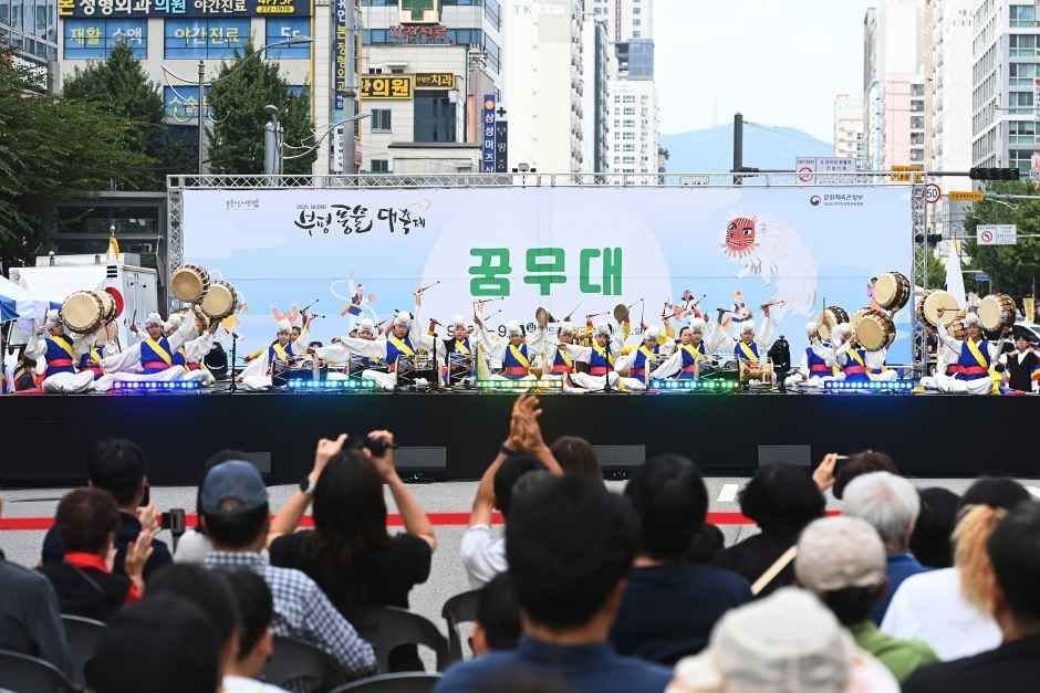 공무대 무대 위에서 풍물패가 북을 치며 공연하고 앞에서 관람객이 사진을 찍는 모습
