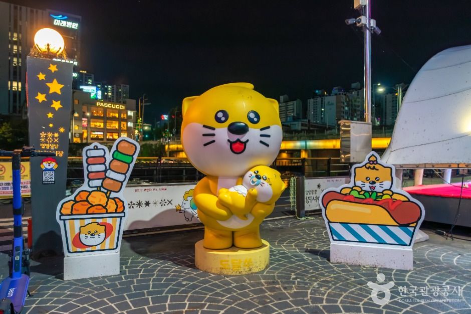 밤 광장에 놓인 떡볶이와 핫도그 모양 입간판과 노란 캐릭터 조형물