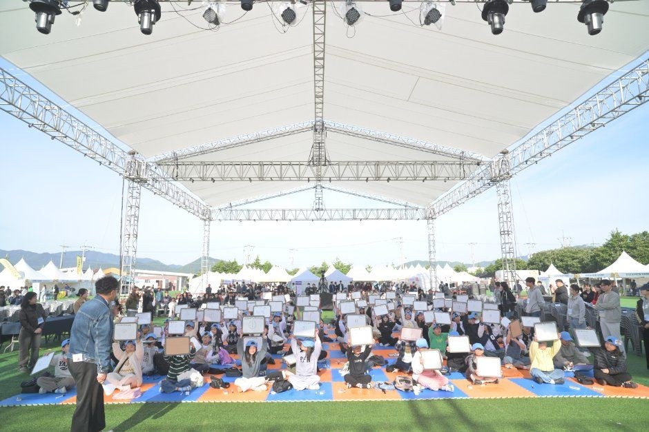 무대 천막 아래 잔디밭에 관람객들이 돗자리에 앉아 행사를 보는 모습