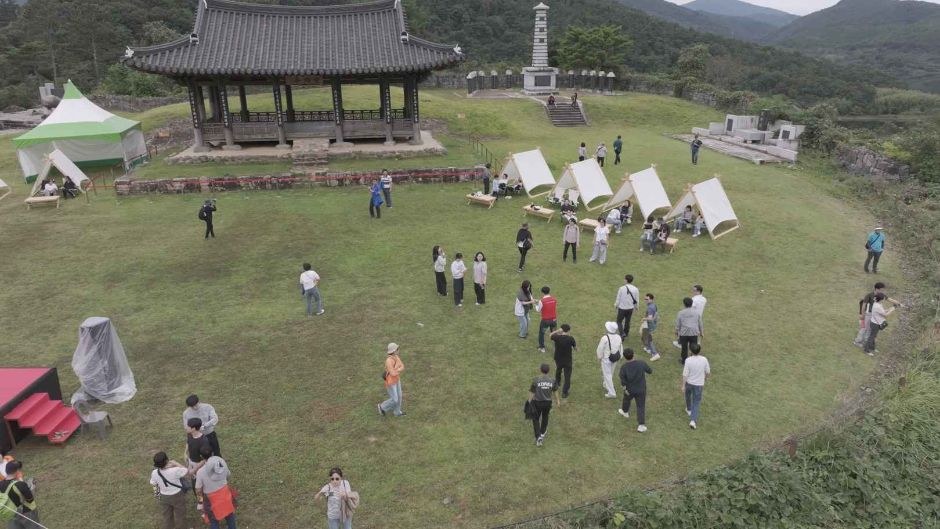 푸른 산을 배경으로 잔디 위에 천막과 정자가 놓이고 사람들이 모인 축제장