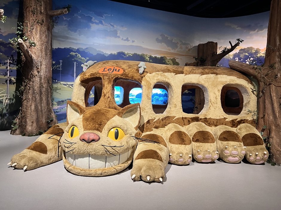 숲과 하늘 배경 앞에 창문마다 영상이 나오는 거대한 인형 고양이버스가 누워 있는 전시 장면