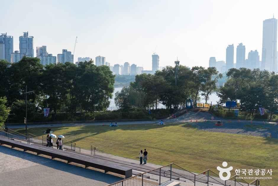 넓은 잔디마당에서 방문객들이 쉬고 있고 멀리 한강과 서울 도심 빌딩숲이 보이는 노들섬 풍경