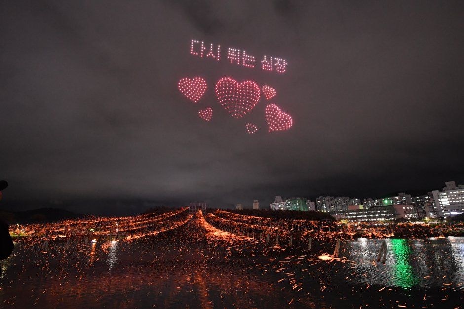 어두운 하늘에 하트 모양을 그린 분홍색 드론 조명이 강물 위에 떠 있는 장면