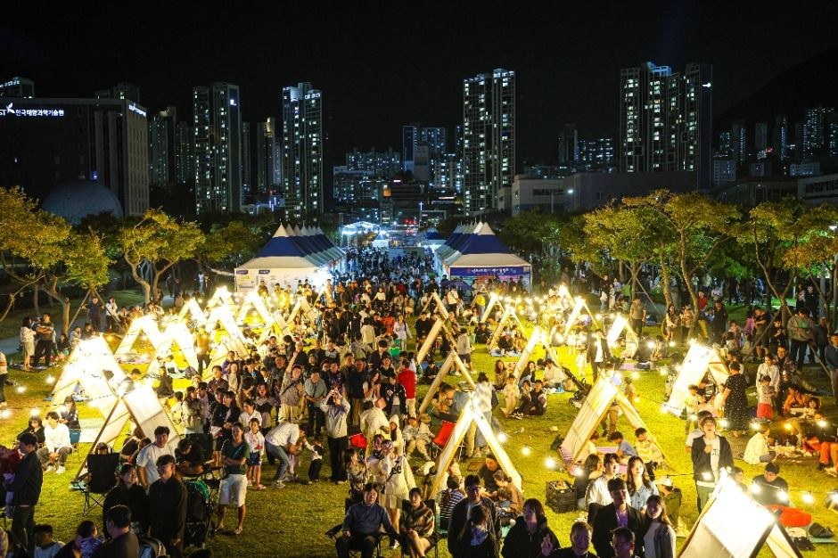 밤에 조명이 켜진 천막과 방문객이 가득한 공원 축제장과 멀리 도시 야경
